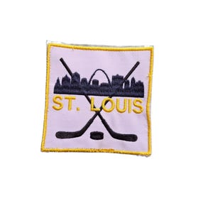 May include: A square embroidered patch with a light purple background and a yellow border. It features a black silhouette of the St. Louis skyline, including the Gateway Arch, crossed black hockey sticks, a black puck, and the yellow text ST. LOUIS.