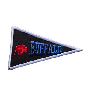 May include: Black pennant-shaped patch with white stitching. Features the word "BUFFALO" in blue embroidered letters and a red bison silhouette. A decorative item for clothing or accessories.