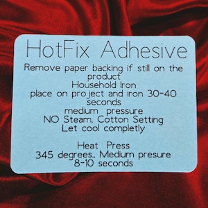 May include: A light blue rectangular card with rounded corners, featuring the text "HotFix Adhesive" and instructions for use with a household iron or heat press. The card is set against a red, textured background.