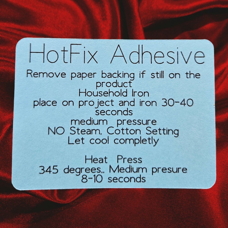May include: A light blue rectangular card with rounded corners, featuring the text "HotFix Adhesive" and instructions for use with a household iron or heat press. The card is set against a red, textured background.