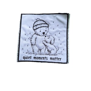 May include: A square white fabric patch with a black embroidered border. The design features a black line art illustration of a bear wearing a beanie and a small dog, with falling snow and the text "quiet moments matter" at the bottom.