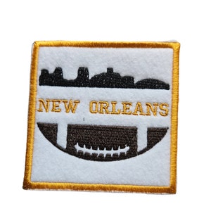 May include: A white embroidered patch with a gold border. The patch features a black silhouette of the New Orleans skyline and a football. The text "NEW ORLEANS" is embroidered in gold below the skyline.