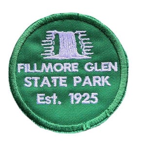 May include: A circular embroidered patch with a green background and a white stitched border. The design features a white waterfall graphic above the text "FILLMORE GLEN STATE PARK" and "Est. 1925" embroidered in white capital letters.