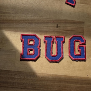 May include: Embroidered patches spelling "BUG" in blue with a red border. The patches are on a light brown wooden surface. The letters are in a bold, block style.