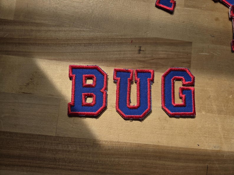 May include: Embroidered patches spelling "BUG" in blue with a red border. The patches are on a light brown wooden surface. The letters are in a bold, block style.