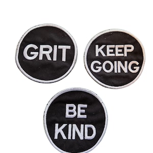 May include: Three round black patches with white text. The patches read "GRIT", "KEEP GOING", and "BE KIND". Each patch has a white border. The patches are likely iron-on or sew-on patches for clothing or accessories.