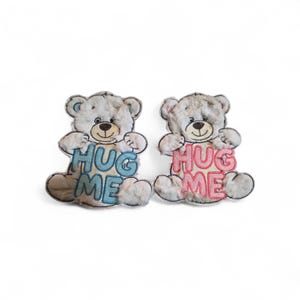 May include: Two plush teddy bears with gray fur and embroidered features. Each bear holds a sign that says "HUG ME" in blue and pink. The bears have a smiling expression and are set against a white background.