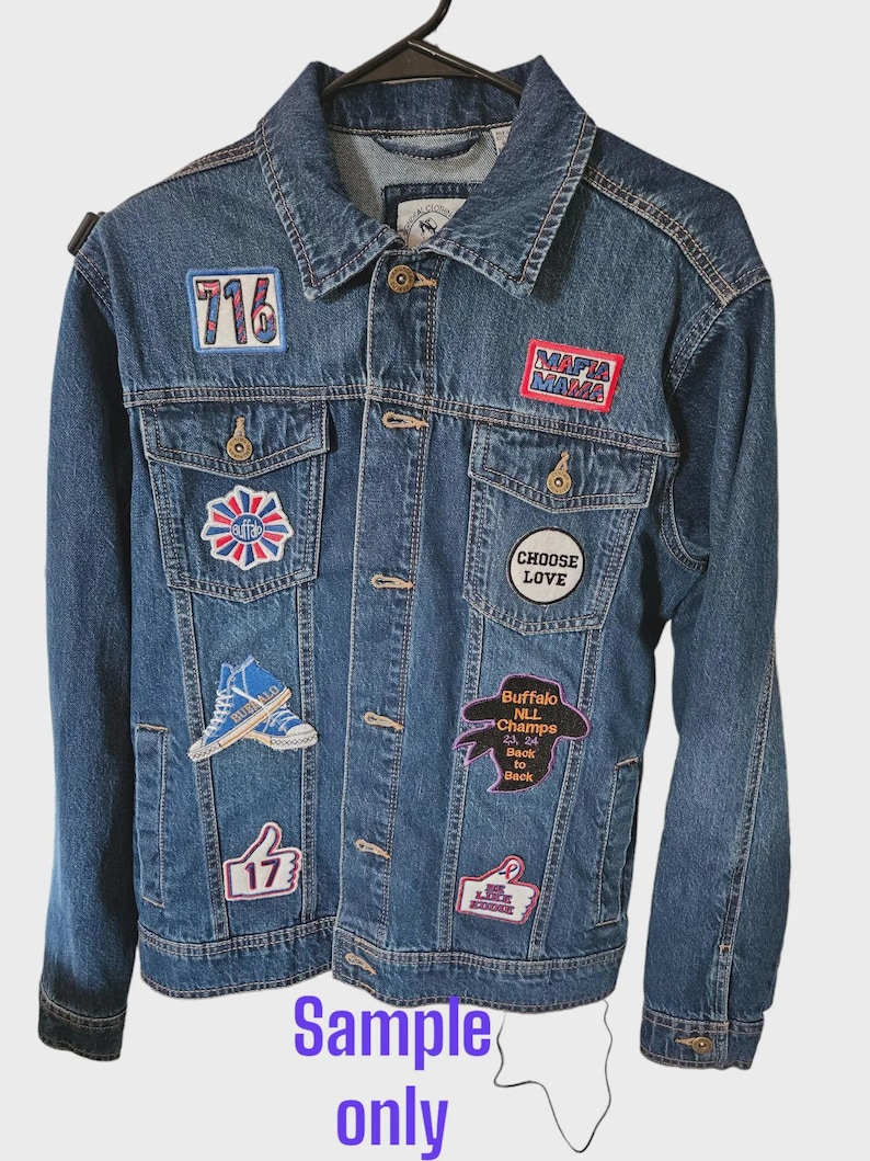 May include: A blue denim jacket adorned with patches displaying text such as "716", "MAFIA MAMA", and "Buffalo NLL Champs 23, 24 Back to Back". The jacket features a traditional design with button fastenings and pockets. The words "Sample only" are at the bottom.