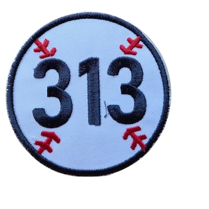 May include: A circular embroidered patch featuring a white background with a dark blue border. The center displays the number 313 in bold dark blue stitching. Four red stitched baseball-style seam accents are positioned around the number.