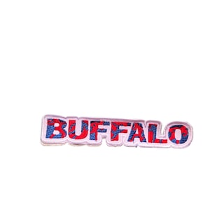 May include: Embroidered patch with the word "BUFFALO" in red letters with blue accents. The patch has a white border and is likely designed to be sewn or ironed onto fabric. The patch is rectangular.