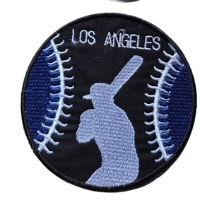 May include: A circular embroidered patch featuring a black background with a light blue silhouette of a baseball player holding a bat. Two white curved baseball stitch patterns flank the player, and the text LOS ANGELES is in white.