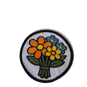 May include: Circular embroidered patch with a floral bouquet design. The flowers are orange, yellow, and blue, with green leaves and a green ribbon. The patch has a black border and a white background.