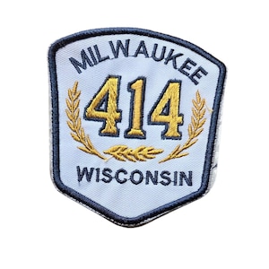 May include: A shield-shaped embroidered patch with a light blue background and a dark blue border. The text MILWAUKEE is at the top, WISCONSIN is at the bottom, and the number 414 is in the center in gold, flanked by two gold wheat stalks.
