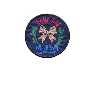 Op de afbeelding: Een ronde donkerblauwe geborduurde patch met een beige strik in het midden, geflankeerd door groene lauriertakken. De tekst "GAME DAY" is in het rood boven de strik geborduurd en "TOUCHDOWN Season" is in het lichtblauw onder de strik.