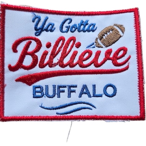 Może przedstawiać: Haftowana naszywka z napisem "Ya Gotta Billieve BUFFALO" w kolorze czerwonym i niebieskim. Brązowa grafika piłki nożnej znajduje się nad słowem "Billieve". Naszywka ma białe tło i czerwoną obwódkę.