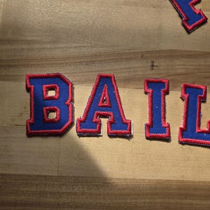 May include: Blue fabric patches spelling "BAIL" in a varsity style font. Each letter has a red outline and border. The patches are on a light brown wooden surface.