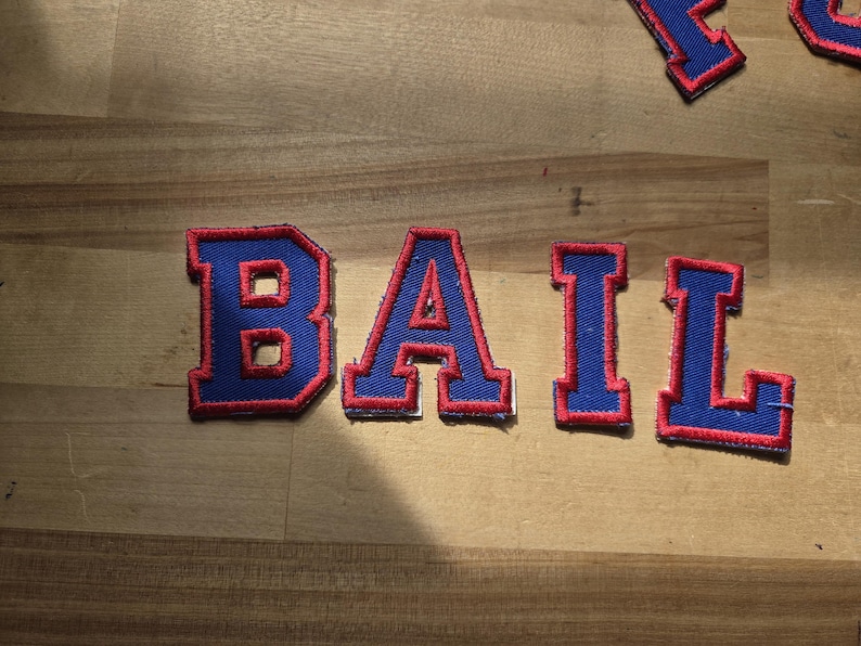 May include: Blue fabric patches spelling "BAIL" in a varsity style font. Each letter has a red outline and border. The patches are on a light brown wooden surface.