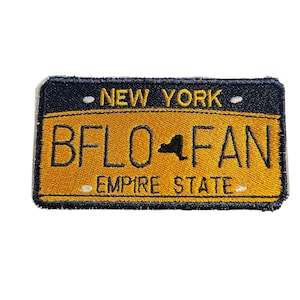 Puede incluir: Un parche bordado amarillo y negro con el texto "NEW YORK" y "BFLO FAN" con una silueta del estado de Nueva York en el medio. El texto "EMPIRE STATE" está debajo del texto "BFLO FAN".