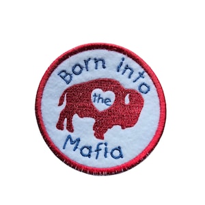 Może przedstawiać: Okrągła, haftowana naszywka z czerwonym wzorem bawołu i niebieskim napisem "Born into the Mafia". Naszywka ma biały środek i czerwoną obwódkę.