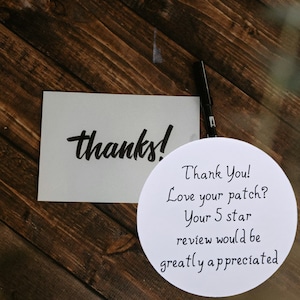 May include: A white card with the word "thanks!" in black script is next to a white circular card with the text "Thank You! Love your patch? Your 5 star review would be greatly appreciated." A black pen is also visible.