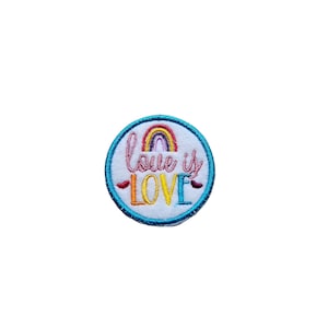 Puede incluir: Un parche redondo azul bordado con un arcoíris y el texto "Love is Love" en rosa y amarillo.
