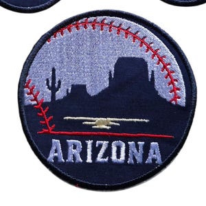 May include: A circular embroidered patch featuring a dark blue silhouette of a desert landscape with a saguaro cactus and rock formations. A red baseball stitch pattern curves across the light blue sky, and the word ARIZONA is in white.