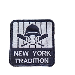May include: A square embroidered patch with a dark navy blue border. The top section features a white baseball, two crossed black baseball bats, and a black baseball cap against a white pinstriped background. The bottom navy blue section reads NEW YORK TRADITION in white text.