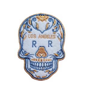以下が含まれることがあります： ライトブルーの背景に、スカルの形をした刺繍パッチ。スカルは金と青の装飾的なアクセント、"LOS ANGELES"の文字と"RR"の文字が金色で施されています。パッチは青い縁取りがされています。