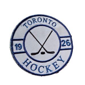 May include: A circular embroidered patch with a white base and blue edging. The patch displays the words "TORONTO HOCKEY" in blue, with crossed hockey sticks and a puck in the centre. The numbers "19" and "26" are also present.