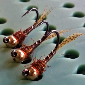 May include: Three brown and gold fly fishing lures with brown and gold feathers. The lures are on a green foam surface.