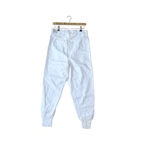 Puede incluir: Pantalón blanco de algodón con cierre de botón y puños elásticos. El pantalón está colgado en una percha de madera con el texto "Arthur Bernard Est. 2012" impreso en ella.