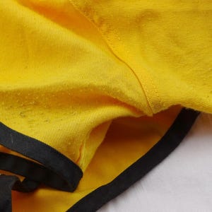 Pu&ograve; includere: Primo piano di pantaloncini gialli con finiture nere. I pantaloncini sono realizzati in un tessuto strutturato e hanno un design semplice e sportivo. Le finiture nere delineano i bordi dei pantaloncini.