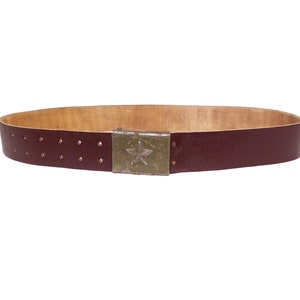 May include: A dark brown leather belt with a metal buckle featuring a five-pointed star. The belt has a tan leather lining and rows of small holes for adjusting the fit.
