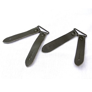 Op de afbeelding: Twee olijfgroene leren riemen met metalen gespen en klinknagels. De riemen zijn rechthoekig met afgeronde uiteinden en zijn verbonden met een metalen lus. Het leer heeft een licht getextureerde uitstraling.