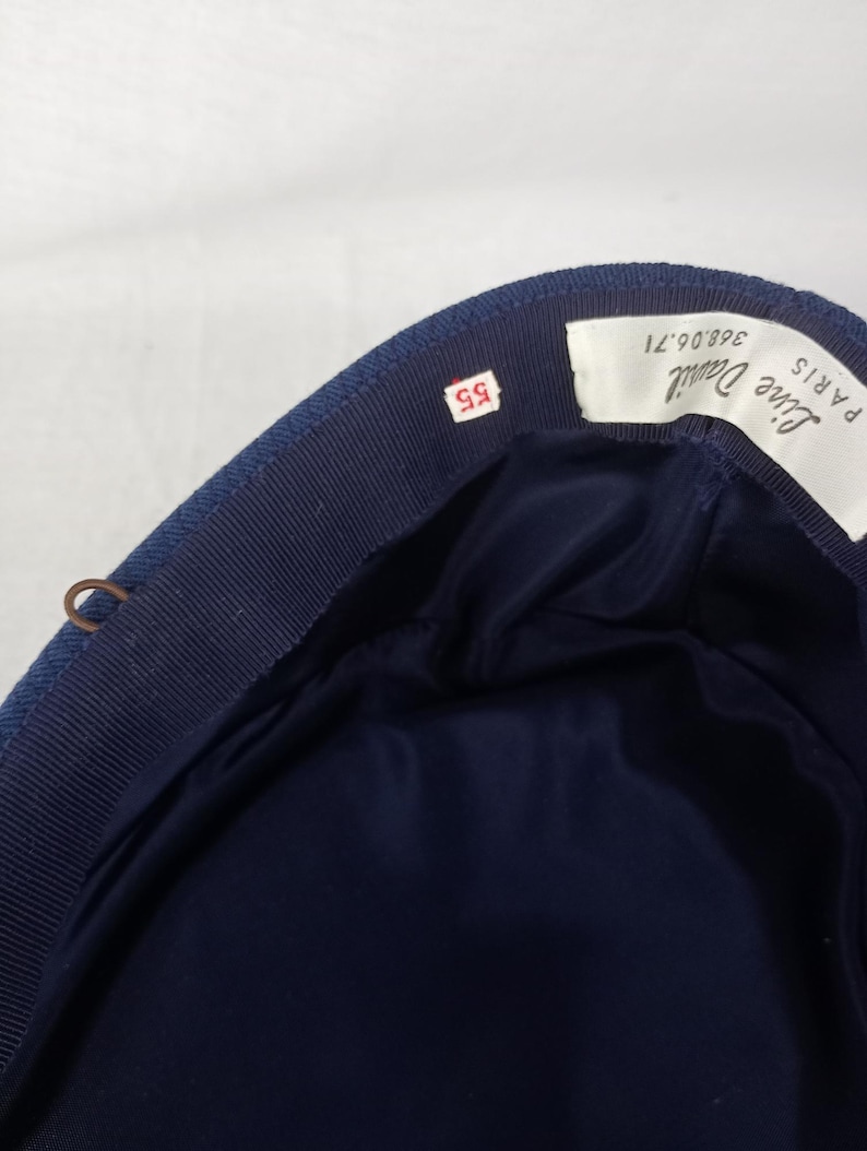 Pu&ograve; includere: Un cappello blu navy con fodera in raso blu scuro. Il cappello ha un bordo in gros-grain e una piccola etichetta con il numero "55". Un'etichetta all'interno recita "Line Daniel PARIS".