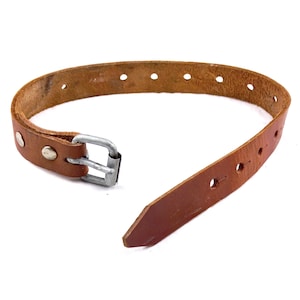 May include: A brown leather strap with a silver metal buckle. The strap has several holes for adjustment and is secured with rivets. The strap is likely for a bag or other accessory.