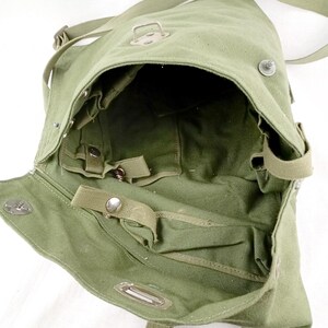 May include: A green canvas military-style shoulder bag with a flap closure and multiple straps and buckles. The bag is open and the interior is visible.
