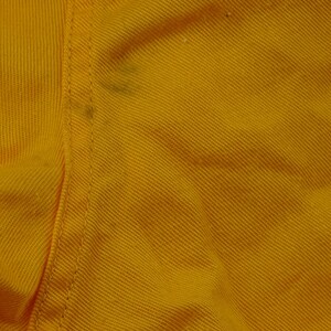 Pu&ograve; includere: Primo piano di una sezione di tessuto giallo con trama diagonale visibile. Una cucitura verticale attraversa il centro, con alcune piccole macchie scure. Il tessuto sembra essere un materiale robusto, forse cotone o una miscela.