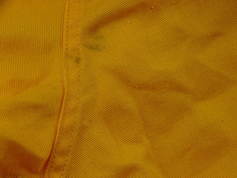 Pu&ograve; includere: Primo piano di una sezione di tessuto giallo con trama diagonale visibile. Una cucitura verticale attraversa il centro, con alcune piccole macchie scure. Il tessuto sembra essere un materiale robusto, forse cotone o una miscela.