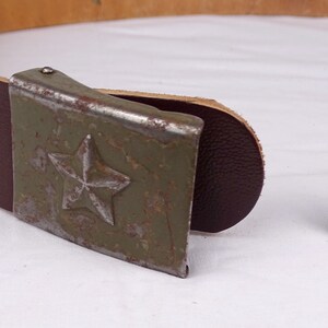 May include: A brown leather belt with a metal buckle featuring a green star. The buckle is worn and has a rustic look.