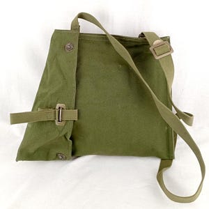 May include: A green canvas bag with a long strap and a buckle closure. The bag has a flap that folds over and is secured with a button.