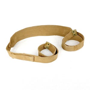 Op de afbeelding: Tan canvas riem met twee lusvormige uiteinden, elk vastgezet met een metalen sluiting. De riem is verstelbaar en heeft een rechthoekige gesp. Het item is waarschijnlijk een onderdeel van een groter stuk apparatuur.