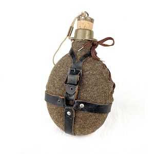 Könnte beinhalten: Eine olivgrüne Vintage-Feldflasche mit Korken, gesichert durch einen schwarzen Ledergurt. Die Feldflasche hat einen braunen Wollbezug und eine braune Kordel am Verschluss. Ein klassisches Militär- oder Outdoor-Ausrüstungsstück.