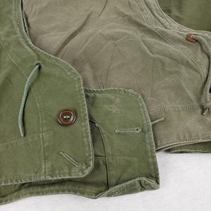 May include: A green canvas jacket with a hood and a button closure. The jacket has a drawstring at the neck and a flap pocket on the chest.