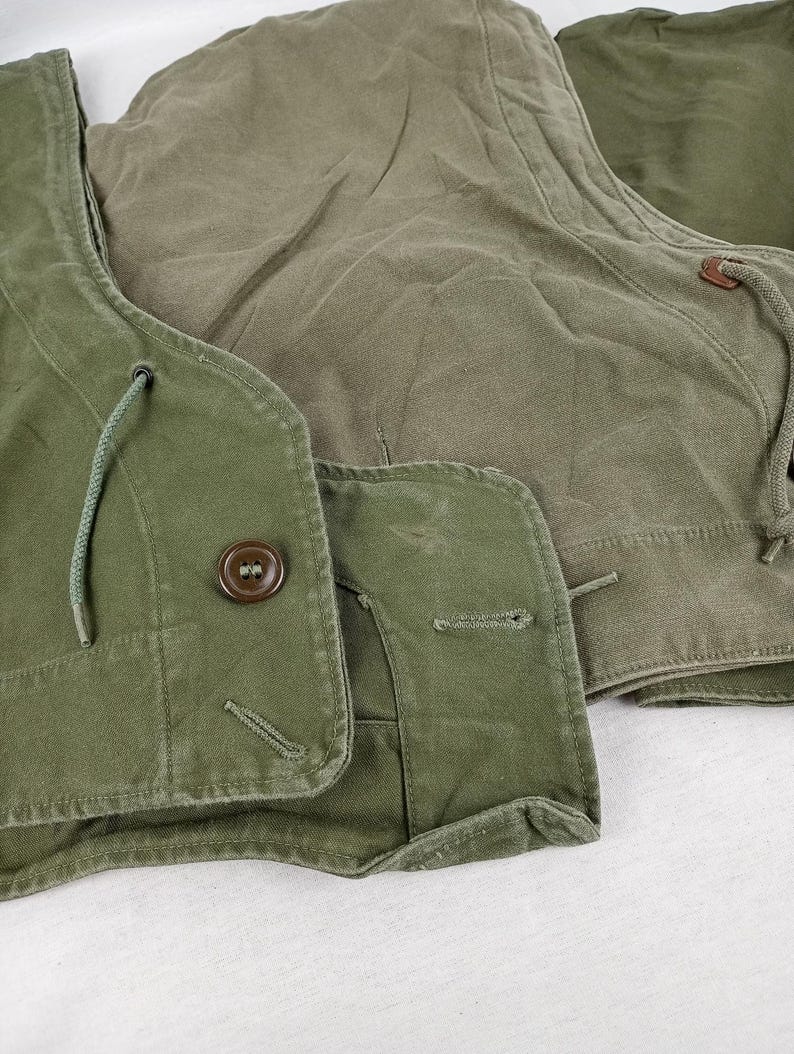 May include: A green canvas jacket with a hood and a button closure. The jacket has a drawstring at the neck and a flap pocket on the chest.