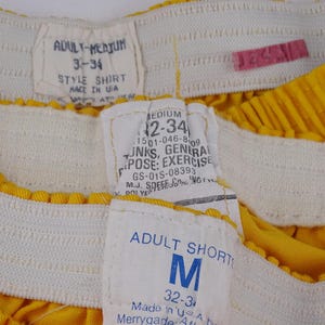 Pu&ograve; includere: Primo piano di pantaloncini sportivi gialli con elastico bianco in vita. I pantaloncini hanno pi&ugrave; etichette con testo tra cui "ADULT-MEDIUM", "STYLE SHIRT", "MEDIUM" e "ADULT SHORTS". I pantaloncini sono realizzati negli Stati Uniti.