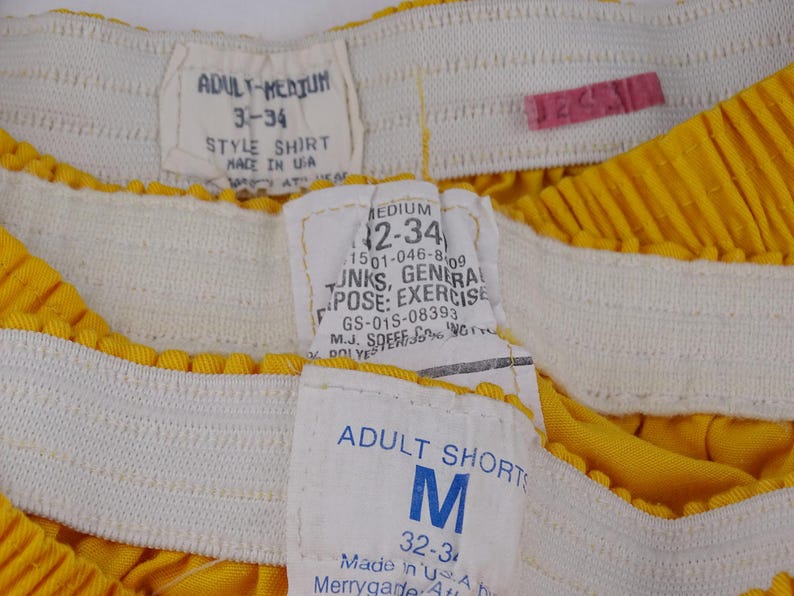 Pu&ograve; includere: Primo piano di pantaloncini sportivi gialli con elastico bianco in vita. I pantaloncini hanno pi&ugrave; etichette con testo tra cui "ADULT-MEDIUM", "STYLE SHIRT", "MEDIUM" e "ADULT SHORTS". I pantaloncini sono realizzati negli Stati Uniti.