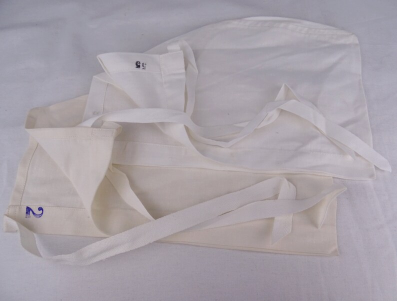 May include: Several off-white fabric caps with attached ties are arranged on a white surface. One cap has the number "2" printed in blue, and another has the number "55" printed in black. The caps are folded and layered.