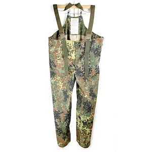 May include: Camouflage pattern overalls with olive green braces. The overalls are made of a durable fabric and feature a button-up front. The camouflage design includes shades of green, brown, and black.