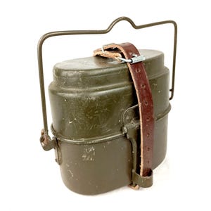 Peut inclure: Un kit de gamelle en métal vert olive de forme rectangulaire arrondie, doté d'une poignée métallique articulée et d'une sangle en cuir marron avec une boucle métallique fixée verticalement au centre pour maintenir les deux parties.